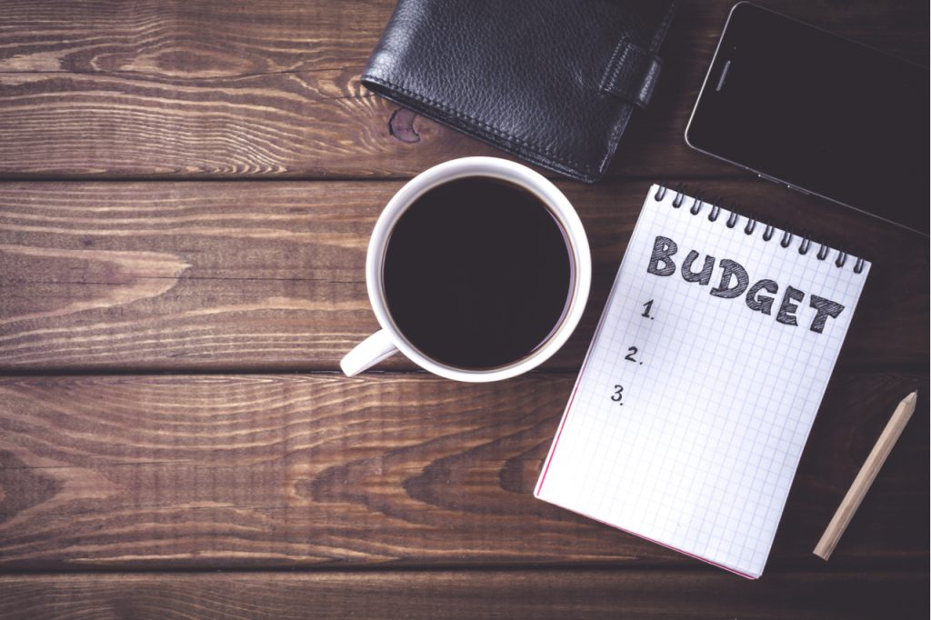 Top view of notepad with word Budget, mobile phone, cup of coffee, pouch on wooden background a start on how to save money.