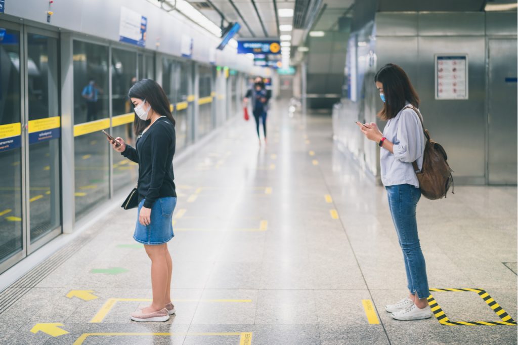 Two asian women wearing protective face mask practicing social distancing while in line waiting for the subway train.