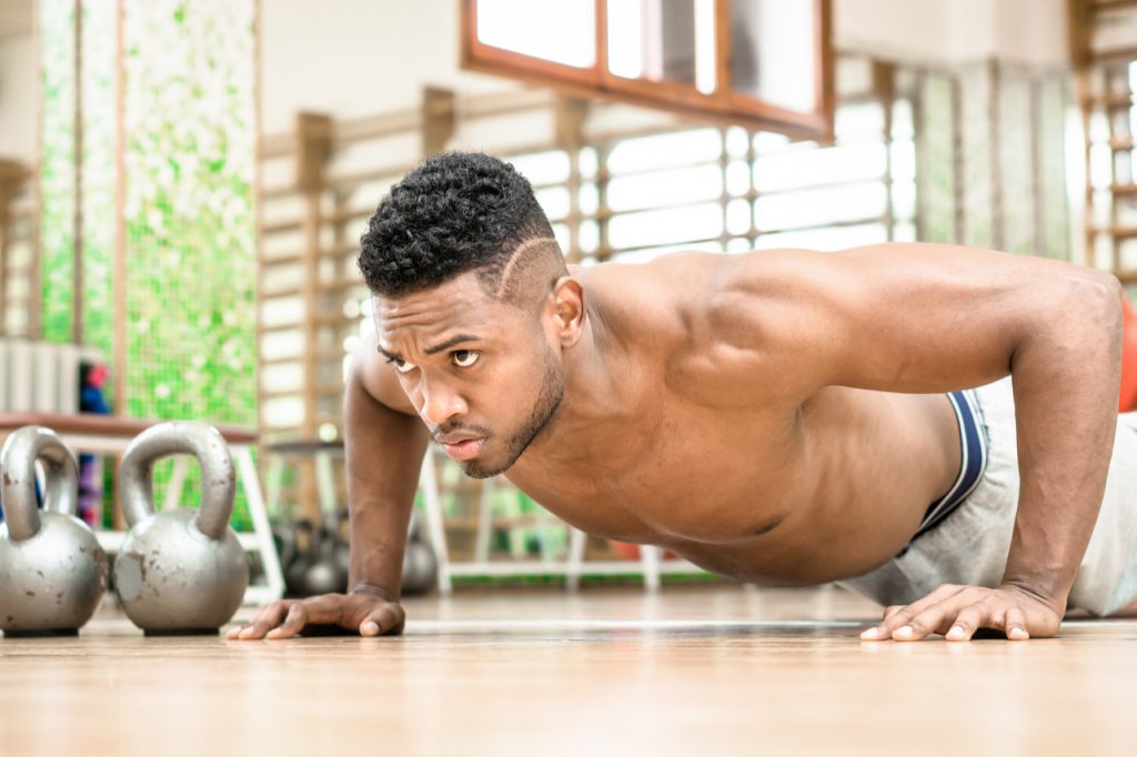A man doing push up working on his bodyweight workout plan with serious facial expression inside gym.