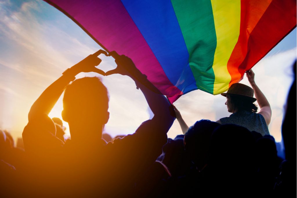 Pride community at a parade with hands raised and the LGBT flag.