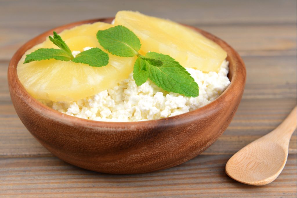 Cottage cheese and 2 slices of pineapple with mint leaves on wooden bowl and a wooden spoon on the side.