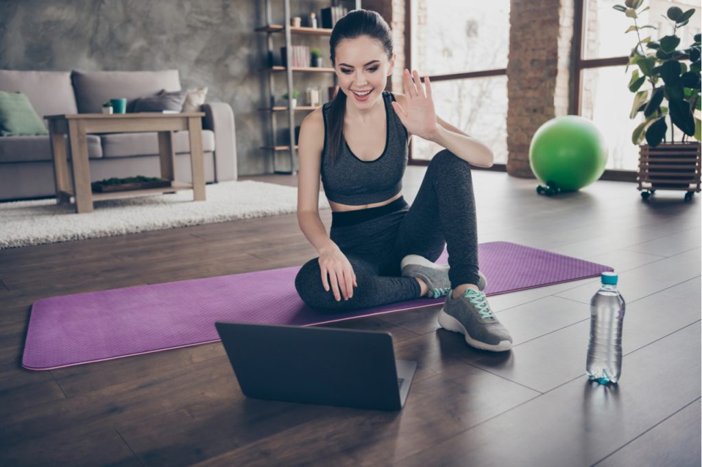 A young woman in sporty clothes sitting on a yoga mat while doing a video call with a friend, celebrating international friendship day.