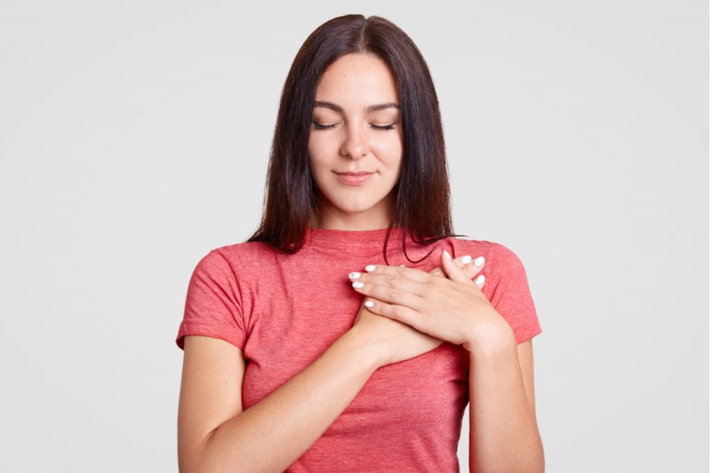 Calm female with closed eyes, keeps both palms on heart, feels gratitude.