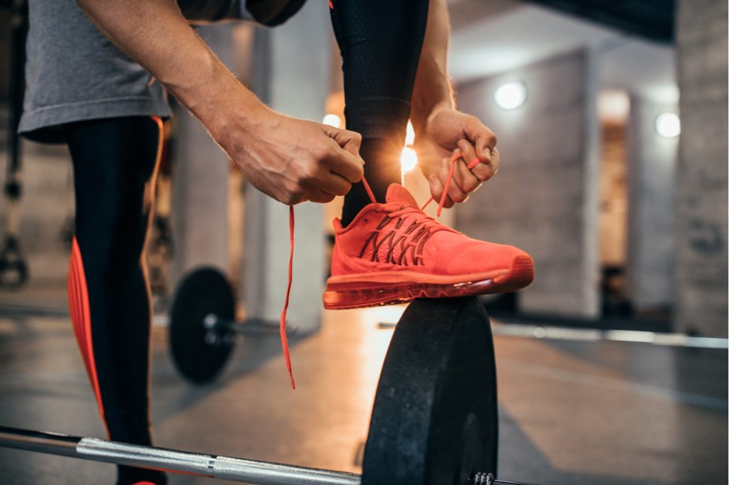 A man tying his shoe laces while stepping on a barbell plate.