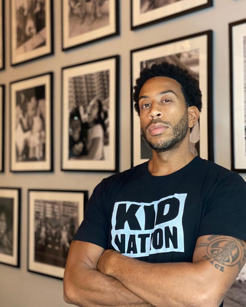 Ludacris wearing a black Kid Nation T-Shirt with black and white photographs in the background.