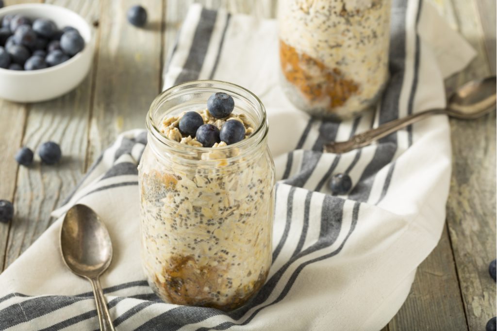 2 jars of overnight oats topped with blue berries placed on top of a napkin and a spoon on the side. 