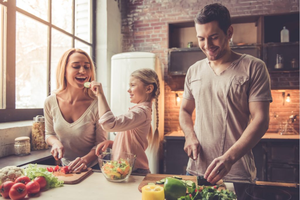 Family of three are cutting vegetables and smiling while cooking in kitchen at home.