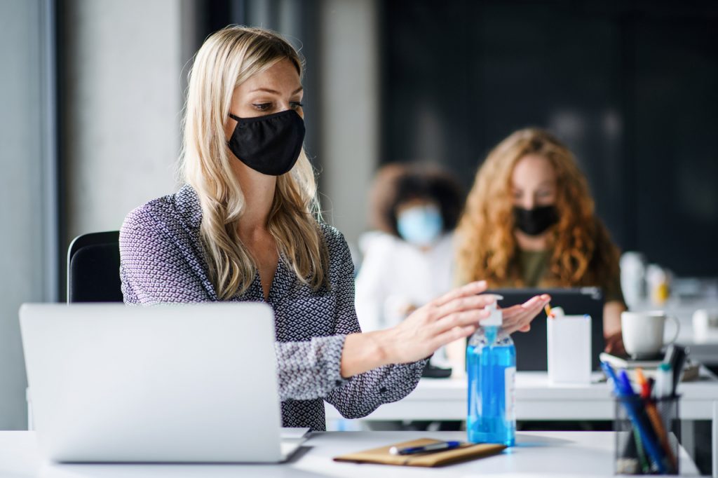 Woman with face mask back at work in office after lockdown, disinfecting hands as part of workplace safety measures.