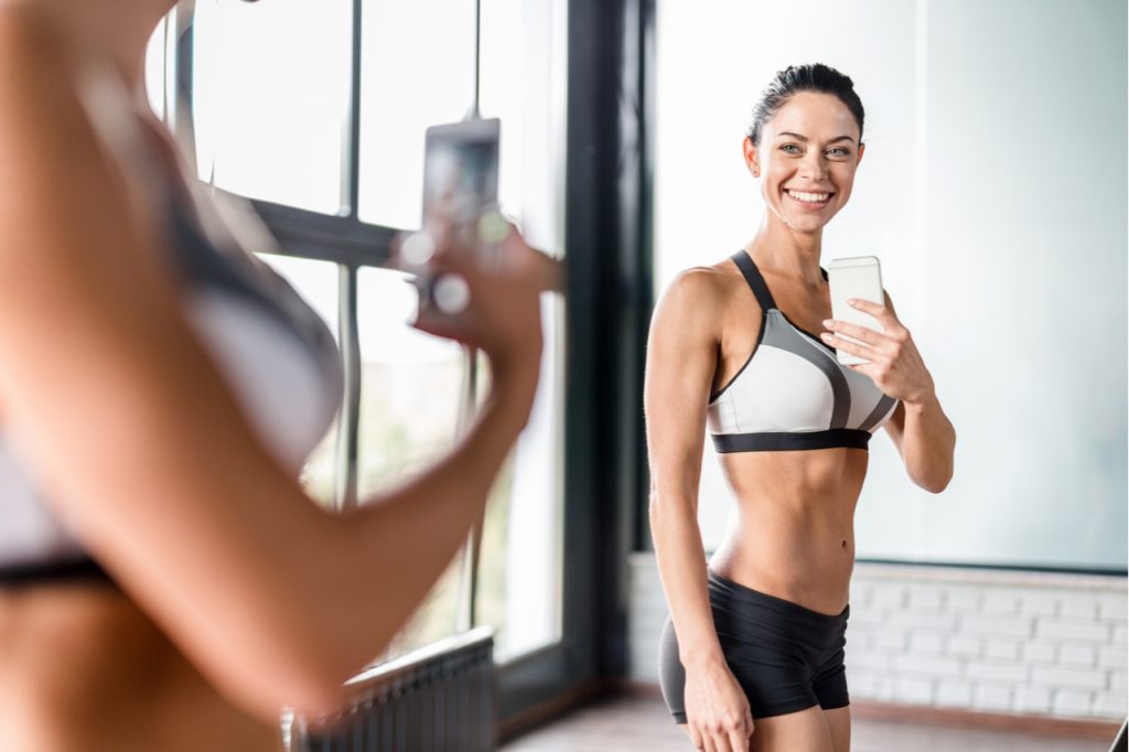 Proud muscular woman smiling boasting her fit figure and slim waist taking selfie in gym mirror.