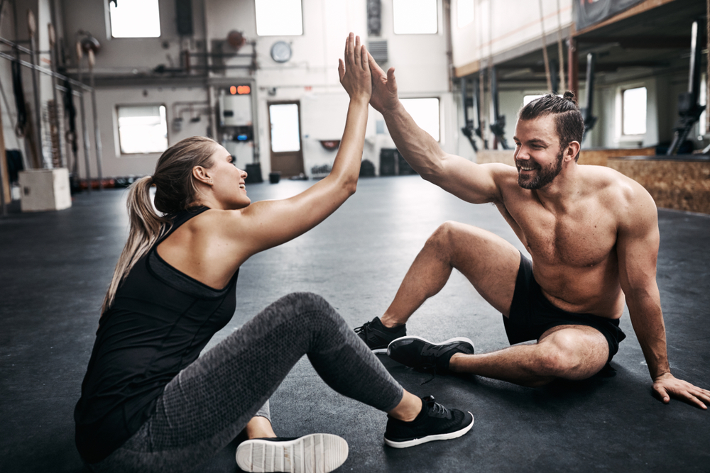 Two fit young people in sportswear smiling and high fiving together while sitting on a gym floor after working out