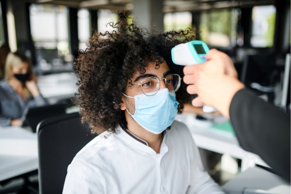 Young man with face mask back at work in office after lockdown, measuring temperature.