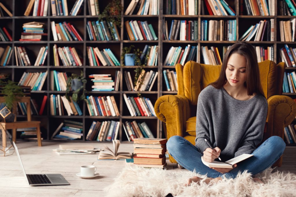 A woman sitting on a carpet while writing on her journal with books on the floor, cup of coffee and a laptop.
