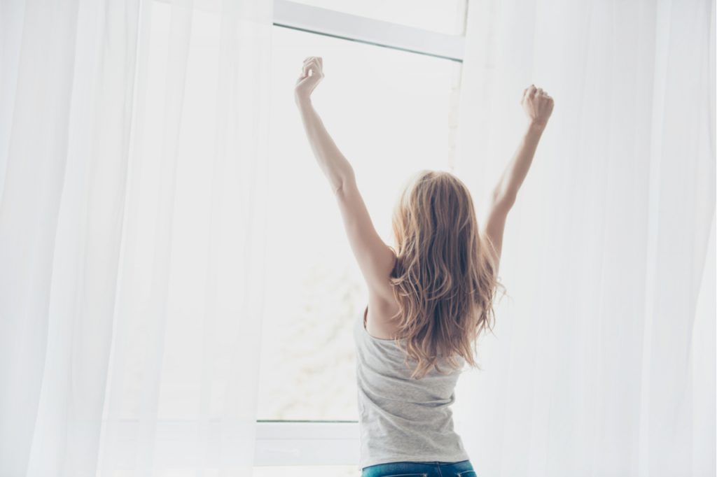 Woman showing positivity after waking up to a beautiful sunshine while standing next to and facing the window.
