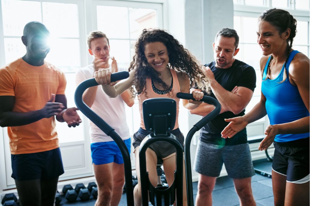 A group of multi racial friends cheering their female buddy while on the stationary bike.