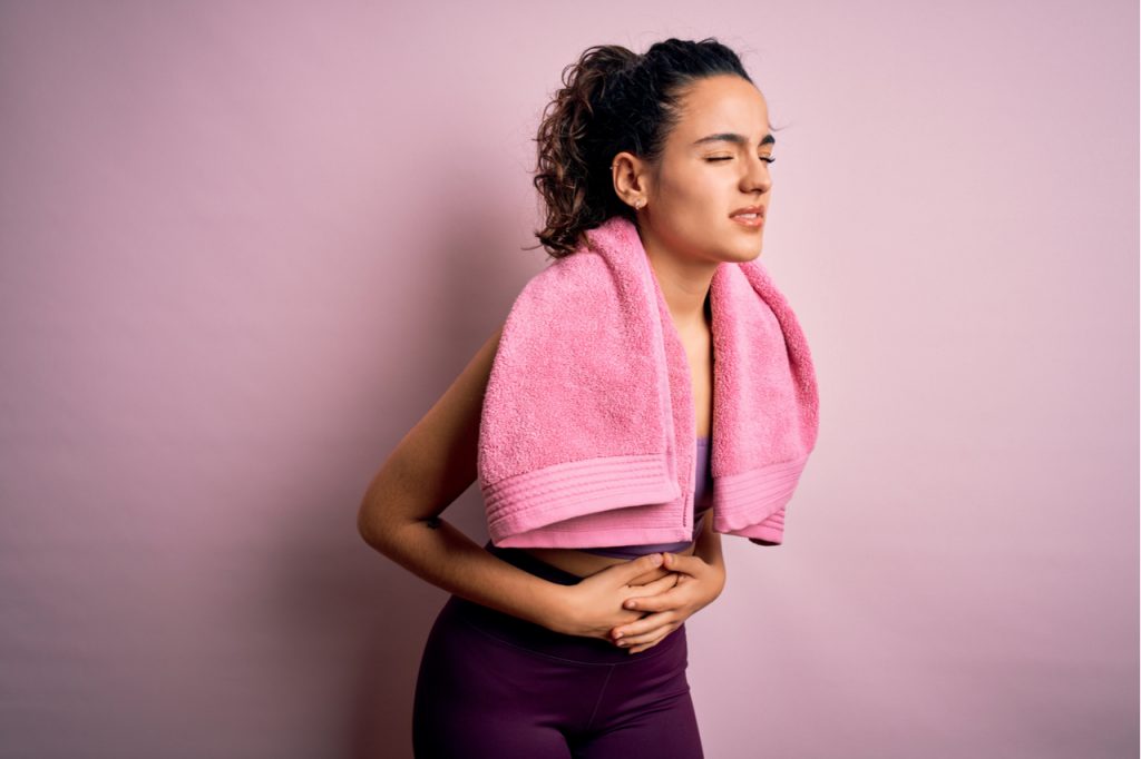 Sportswoman with curly hair with towel over pink background with hand on stomach because stomach pain.