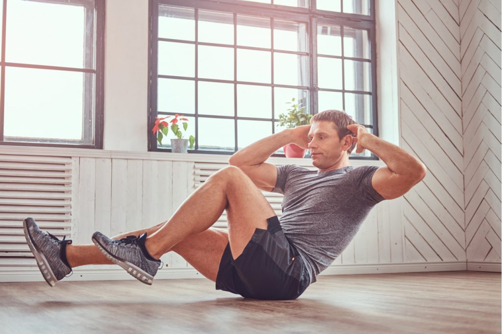 Man in a t-shirt and shorts doing abdominal exercises on floor at home promoted by shine text.