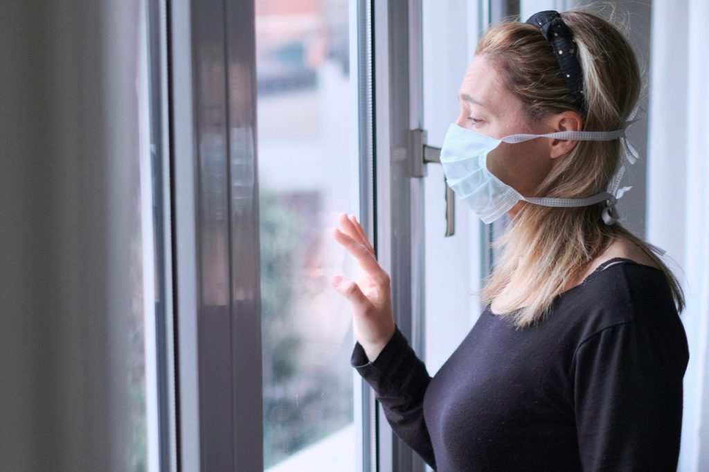 A woman wearing a mask looking through the window while at home looking stressed which is one of the few covid-19 emotions.