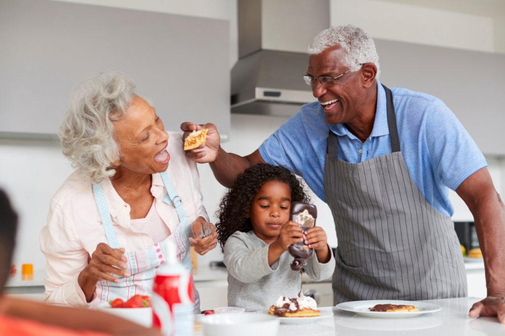 Grandparents In Kitchen With Granddaughter Making Pancakes Together.
