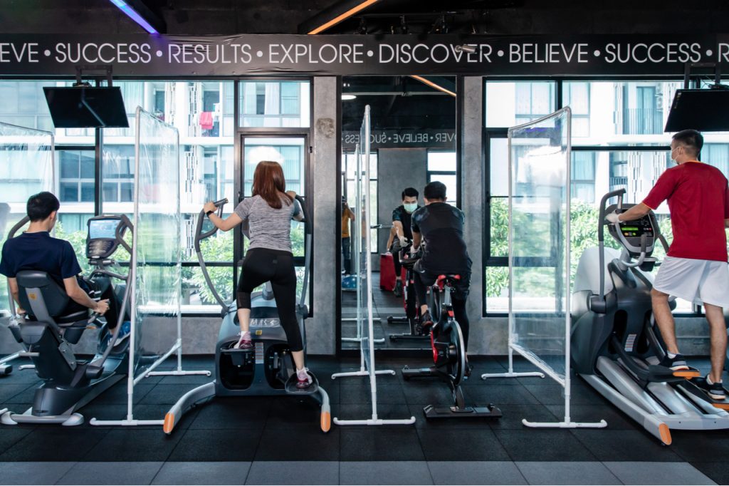 A few people working out at the gym. The gym uses barriers in between gym equipment to ensure safety and social distancing.