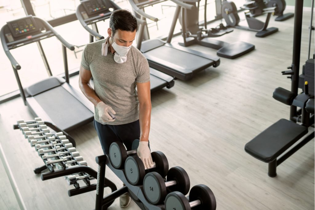 A man wearing a face mask wiping and disinfecting gym equipment.