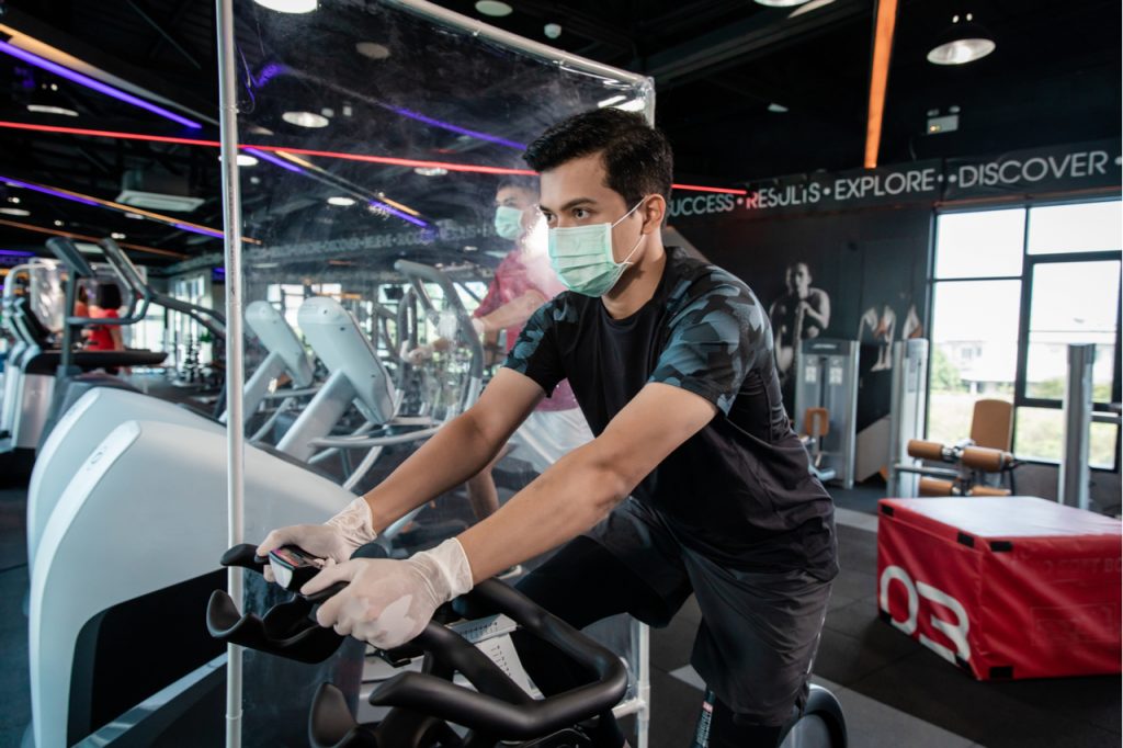 Asian man working out on a stationary bike at the gym while wearing a mask. The gym uses plastic barriers in between workout equipment as part of the new normal.