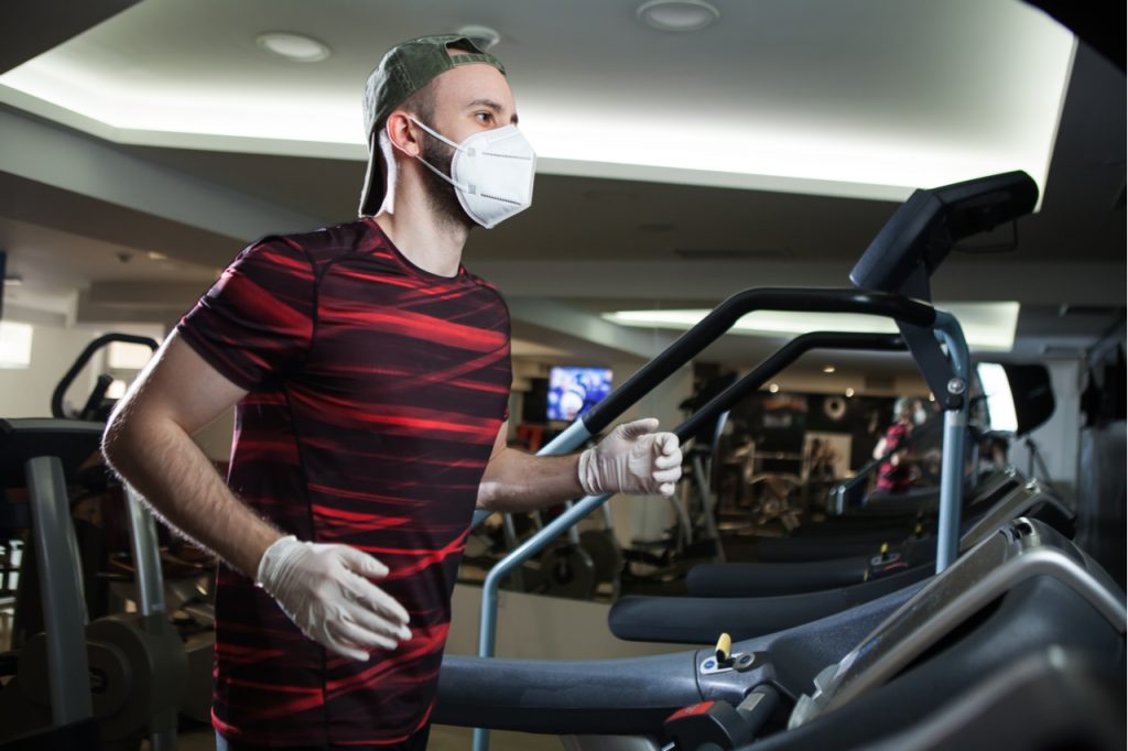 A man wearing gloves and a face mask running on a treadmill at the gym.