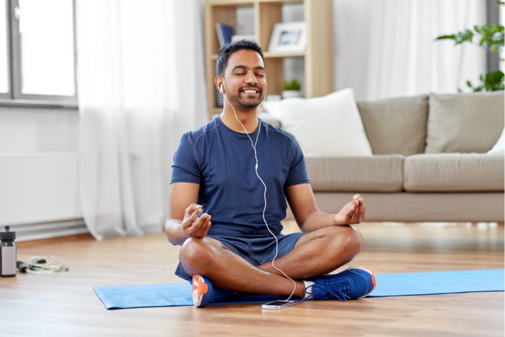Man in earphones listening to music on smartphone and meditating in lotus pose at home for positive energy.