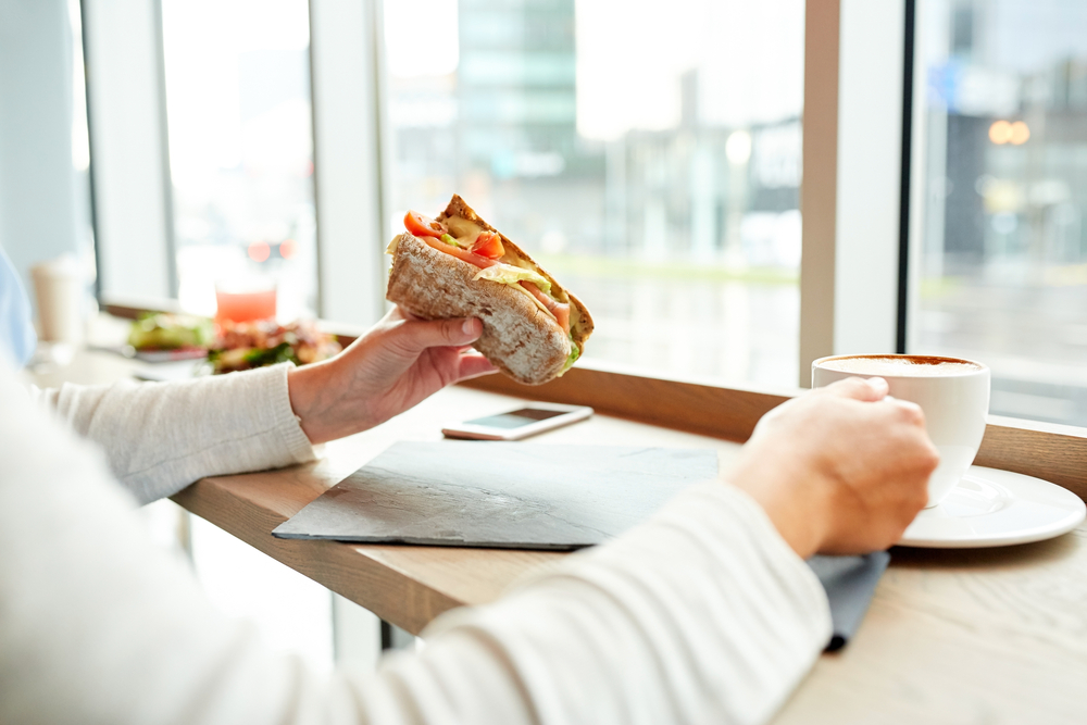 woman eating salmon panini sandwich with tomatoes and cheese and drinking coffee for lunch at restaurant