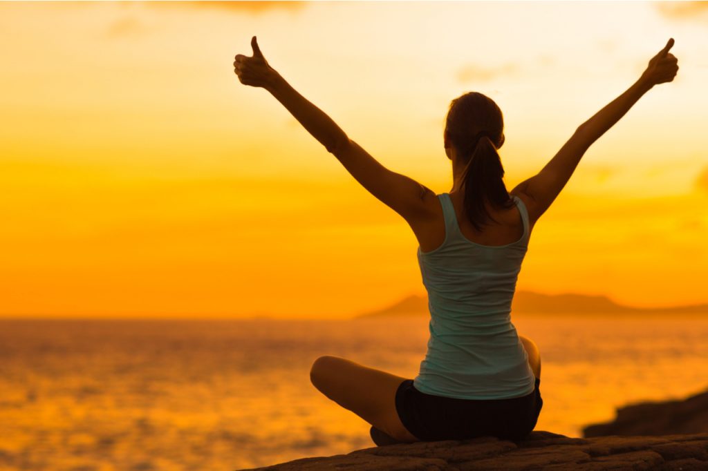 A woman sitting on a rock facing the sea while here hands are up in the air.