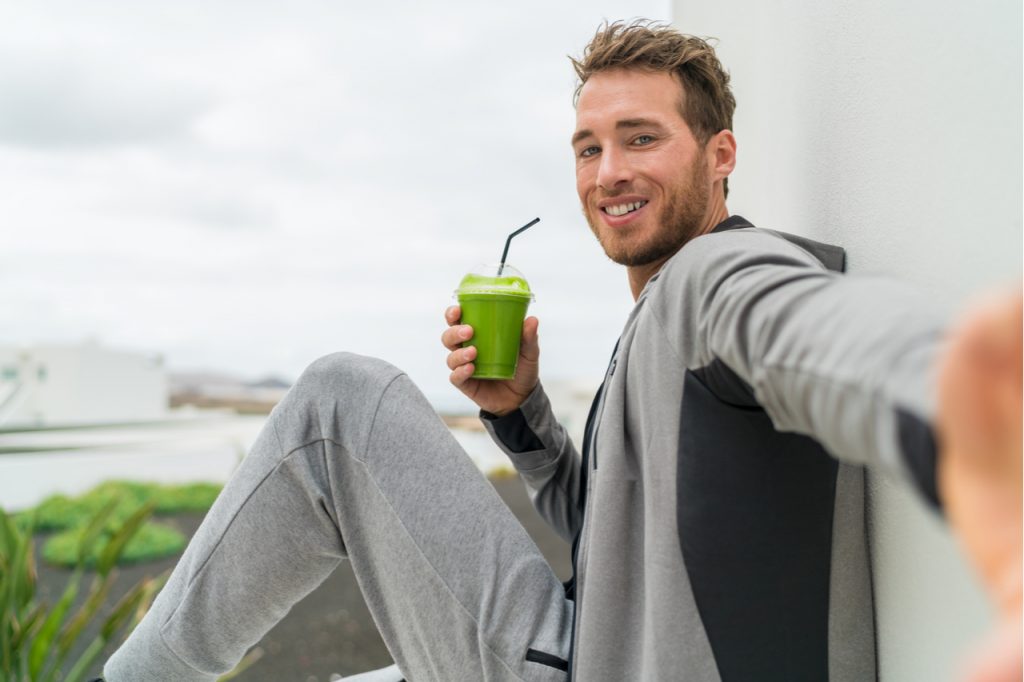 Fitness man taking self portrait picture at gym drinking vegetable juice after workout in sweatpants and sportswear.