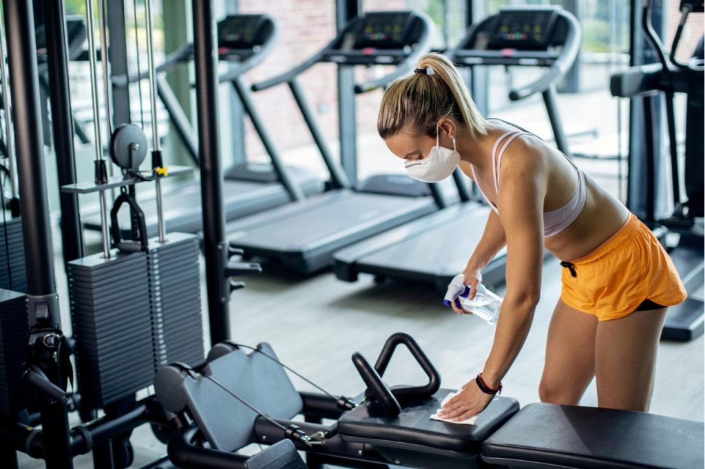 A fit woman disinfecting a gym equipment while wearing a mask.