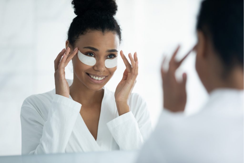 An Afro-american women applying collagen eye patch while in front of the mirror.