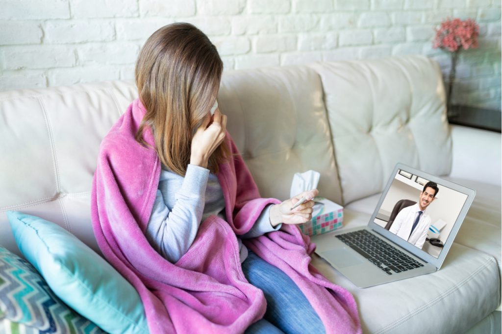 A woman wrap in a blanket taking her temperature while talking to her doctor online. 