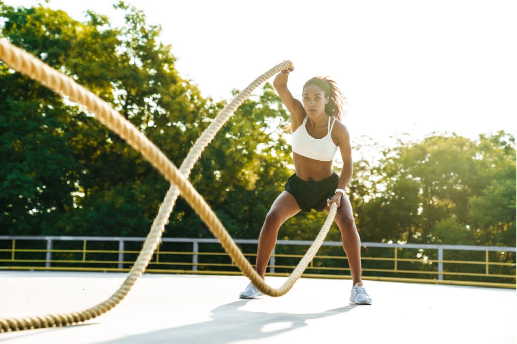 Woman in sportswear doing workout with battle ropes at playground outdoors.