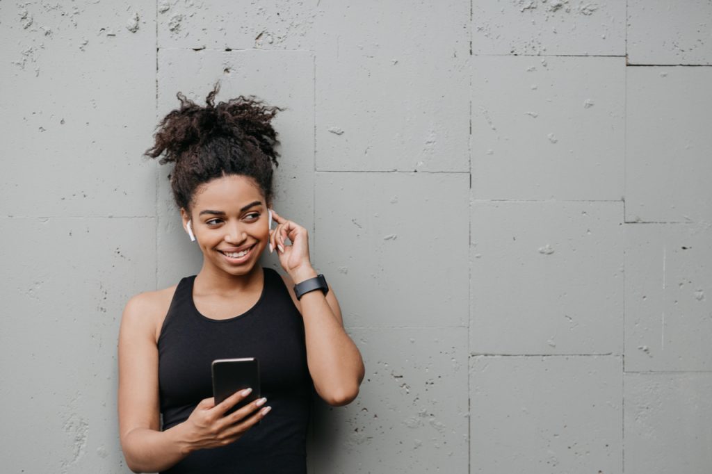 Smiling african american girl in sportswear puts on wireless headphones and holds smartphone in hand.