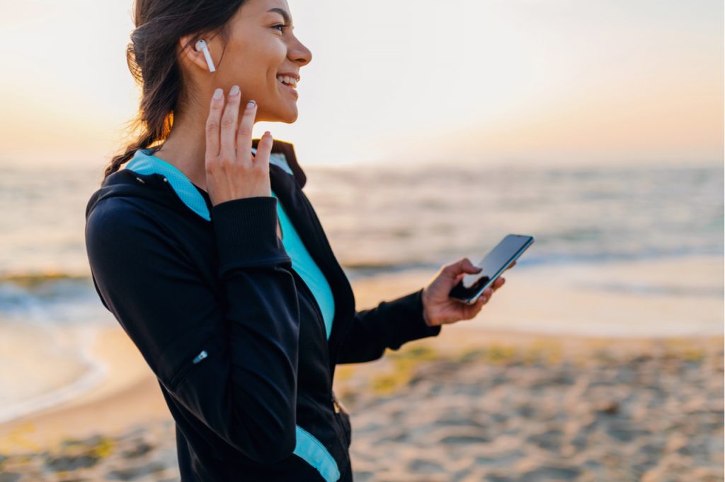 Woman doing sport exercises on morning sunrise beach in sports wear listening to music on wireless earphones holding smartphone.