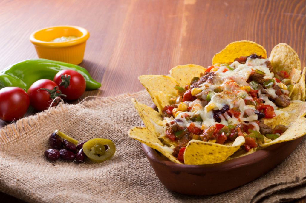 One of the best nacho recipe is chicken Nachos. Baked in casserole with cheese, beans, corn, hot jalapeno, chicken, red pepper and mushrooms serving on wood table.