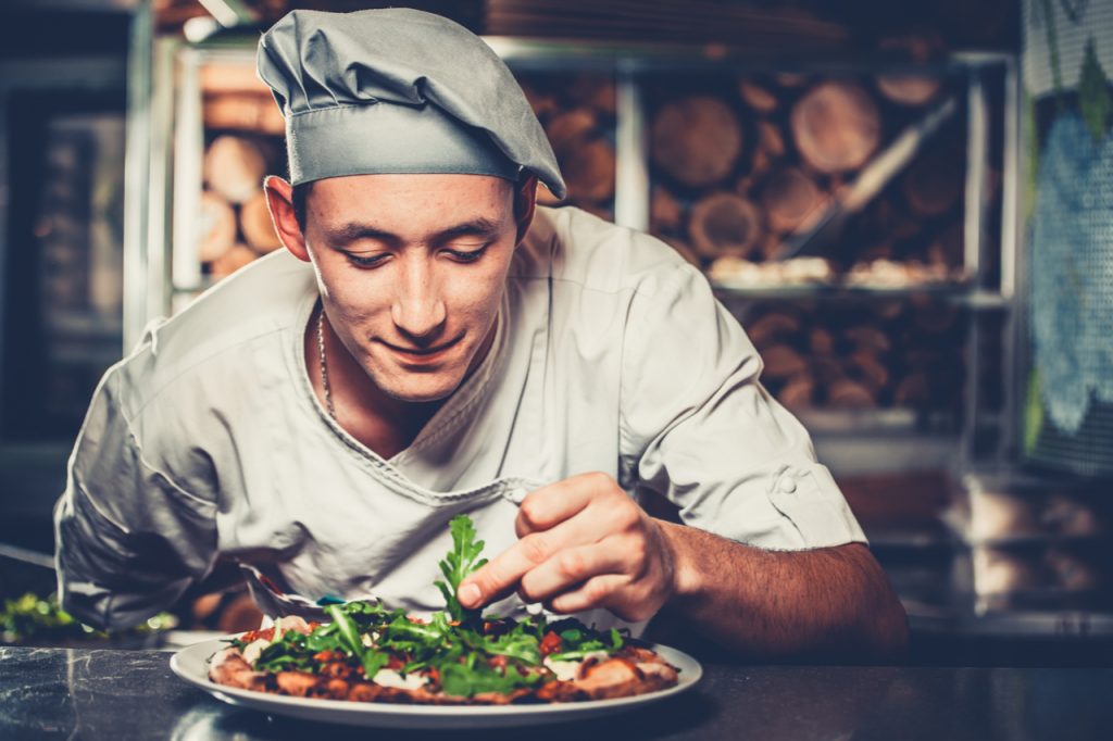 Young smiling chef in white uniform and gray hat decorate ready dish with green rucola herbs in interior of modern restaurant kitchen.