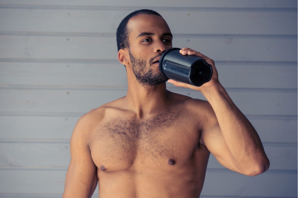 Handsome Afro American sportsman is drinking protein shake while standing topless in front of gray wall.