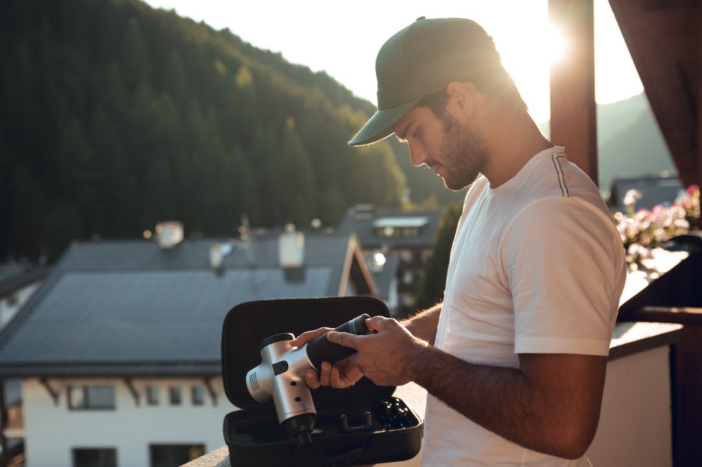 Man with cap and white shirt holding the sonic x massage gun his arm and body at sunset at home.