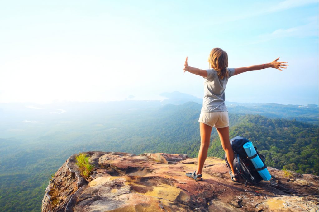 Woman with backpack standing on a rock with raised hands and looking to a valley below.