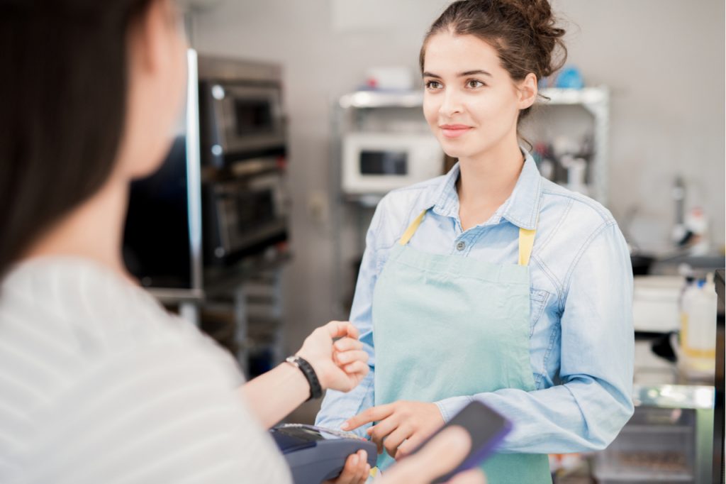 Waist up of smiling young woman wearing apron looking at customer paying via garmin vivoactive 3.