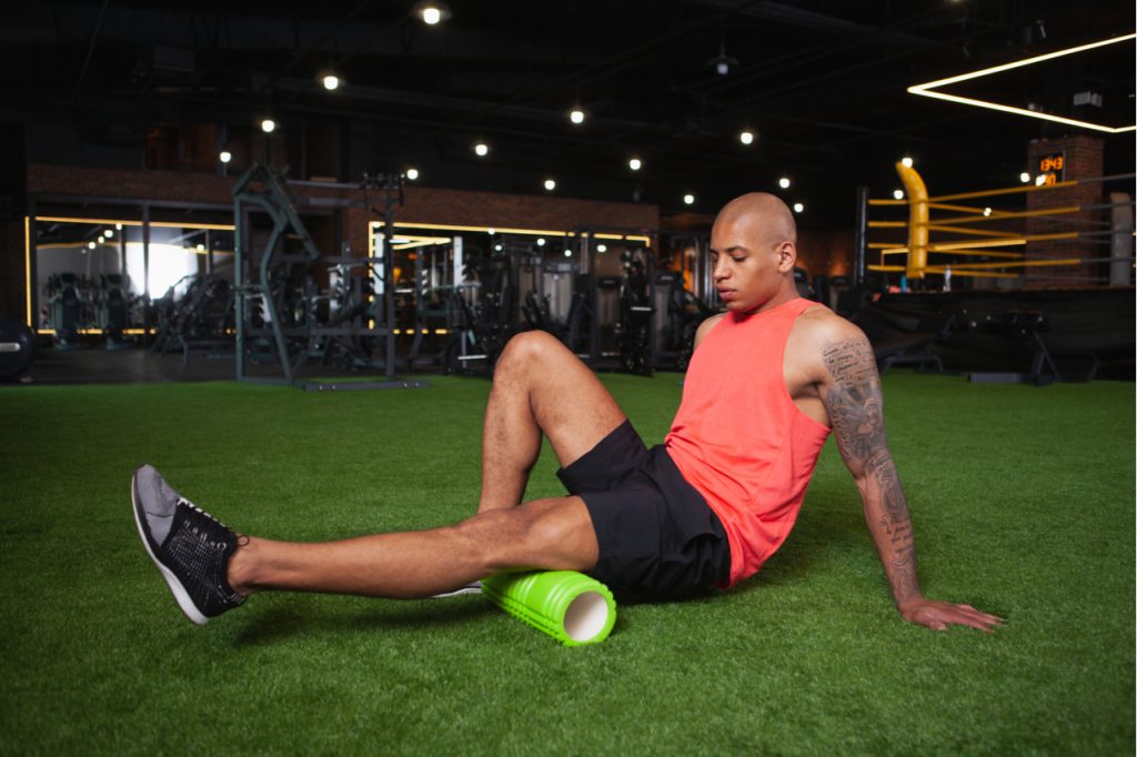 Male athlete relaxing leg muscles after gym workout, using foam roller.