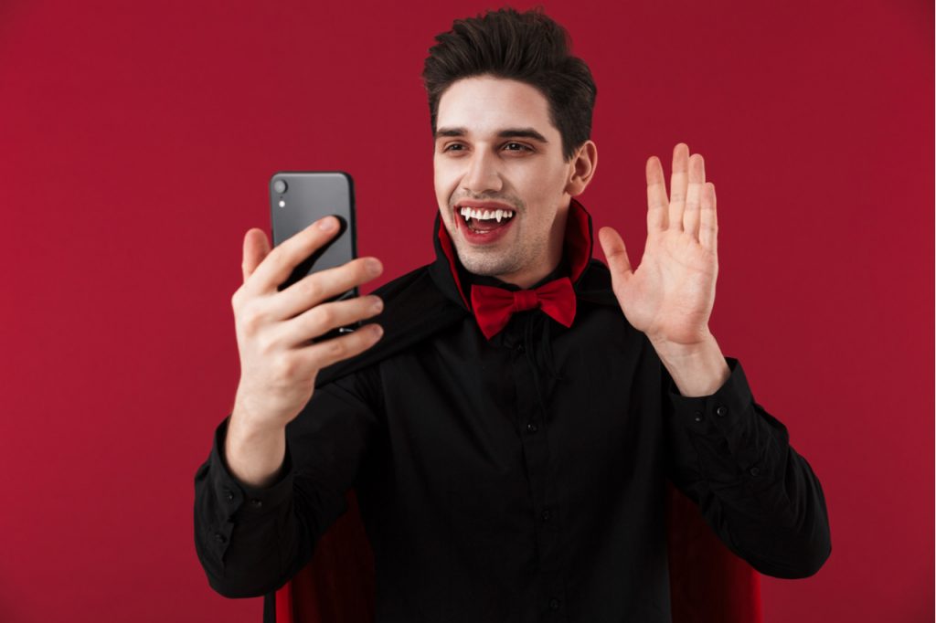 Image of vampire man with blood and fangs in black Halloween costume looking at smartphone isolated over red wall.