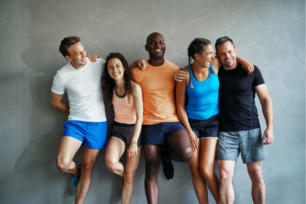 Smiling group of friends in sportswear laughing together with positive mindset while standing arm in arm in a gym after a workout.