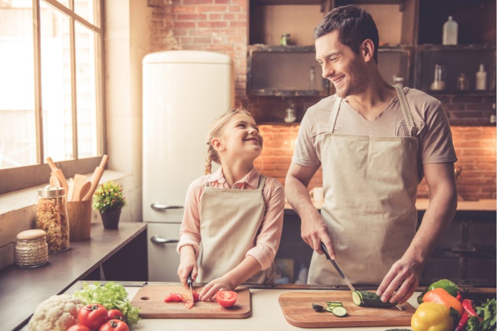 Dad and daughter are cutting vegetables and smiling while cooking in kitchen at home.