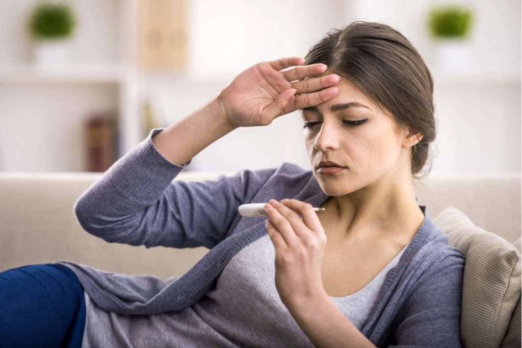 A woman looking at a thermometer while feeling her head for fever.