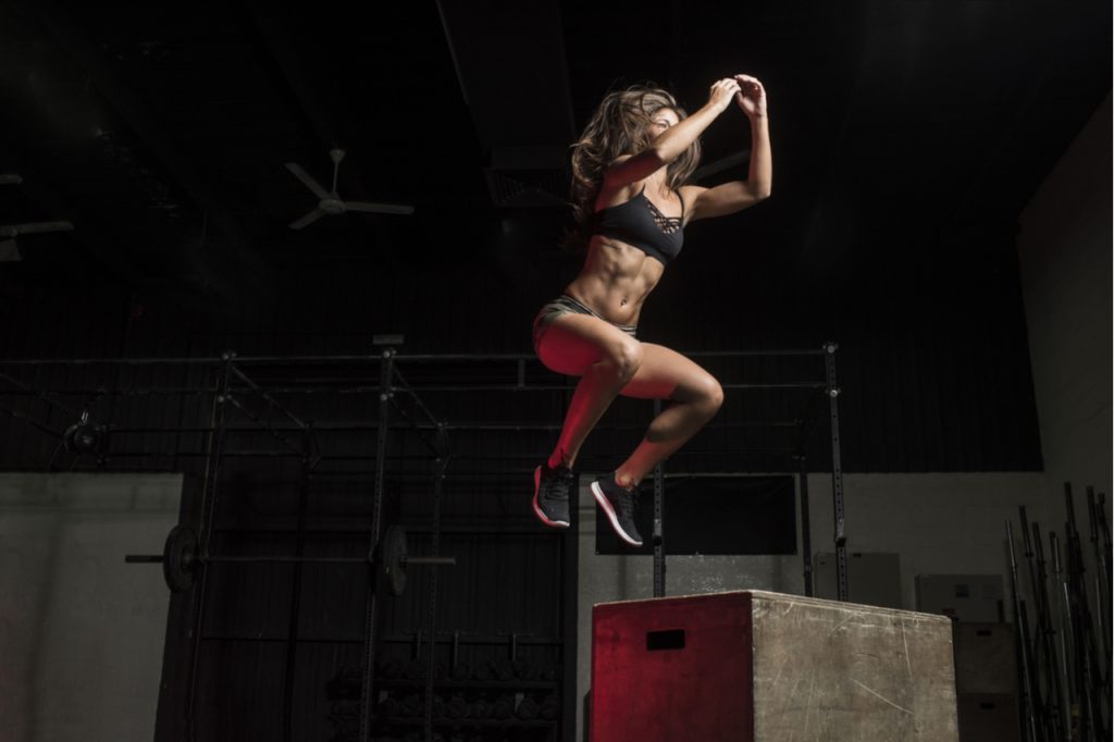 Fitness athlete performs box jumps in a dark gym wearing black sports top and short tights.