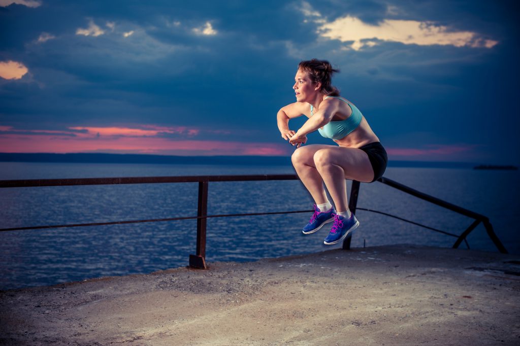 Young strong woman in sportswear doing plyometric exercises on pier. Jump squats, fitness workout outdoors.