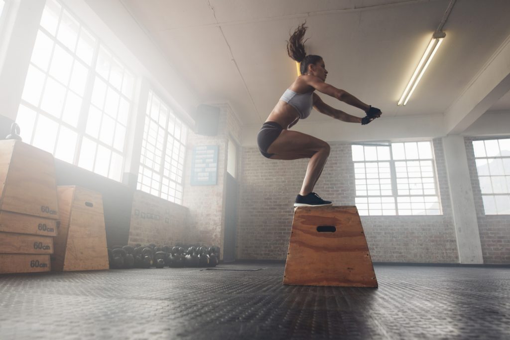 Side view image of fit young woman doing a box jump exercise. Muscular woman doing a box squat at the gym.